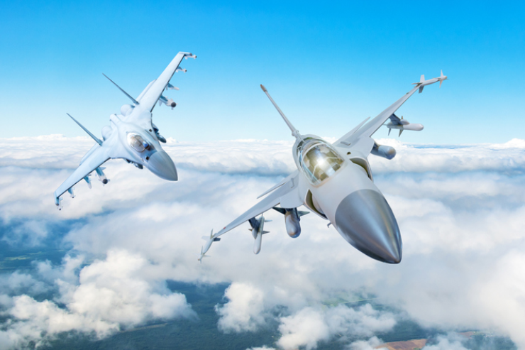 Two fighter jets soar above white clouds in a bright blue sky, banking sharply with power and agility.
