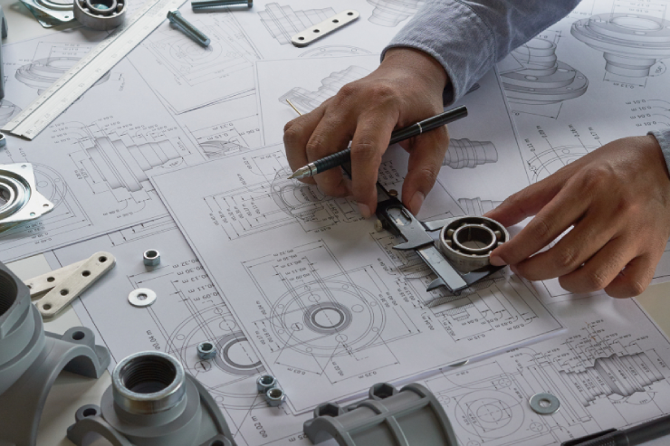 A person’s hands work on technical drawings, using a caliper to measure a mechanical part. Designs and engineering tools are scattered across the table.